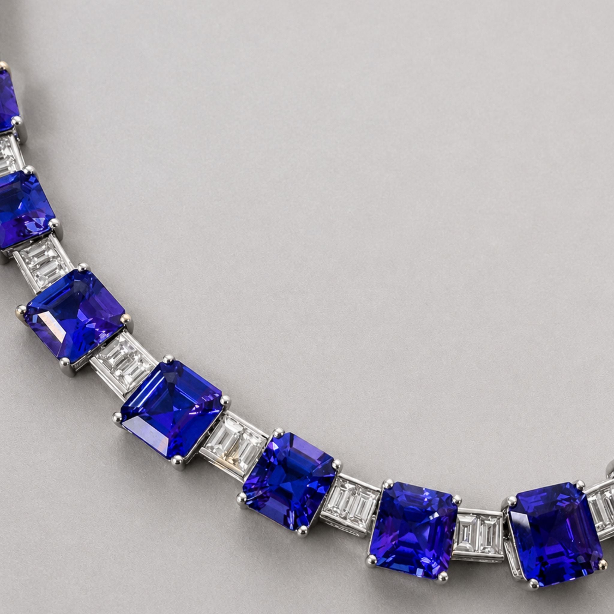 A close-up of necklace featuring large, deep blue emerald-cut tanzanites set with baguette-cut diamonds stones, photographed on a neutral gray background.