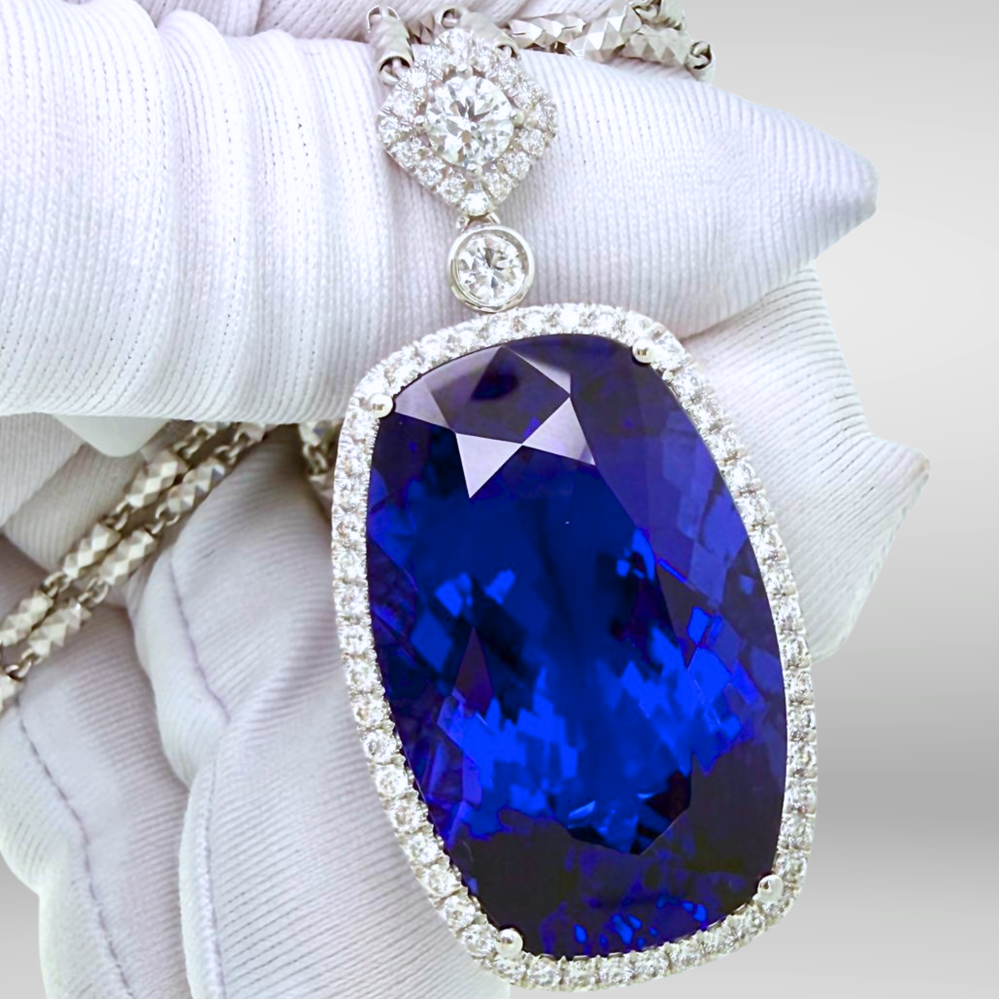 A close-up of a white-gloved hand holding a large oval tanzanite investment grade 50 carat pendant necklace. The pendant features a vivid deep blue faceted vb 6/6 tanzanite gemstone,  surrounded by a halo of diamonds. For sale at tanzanite direct. , 