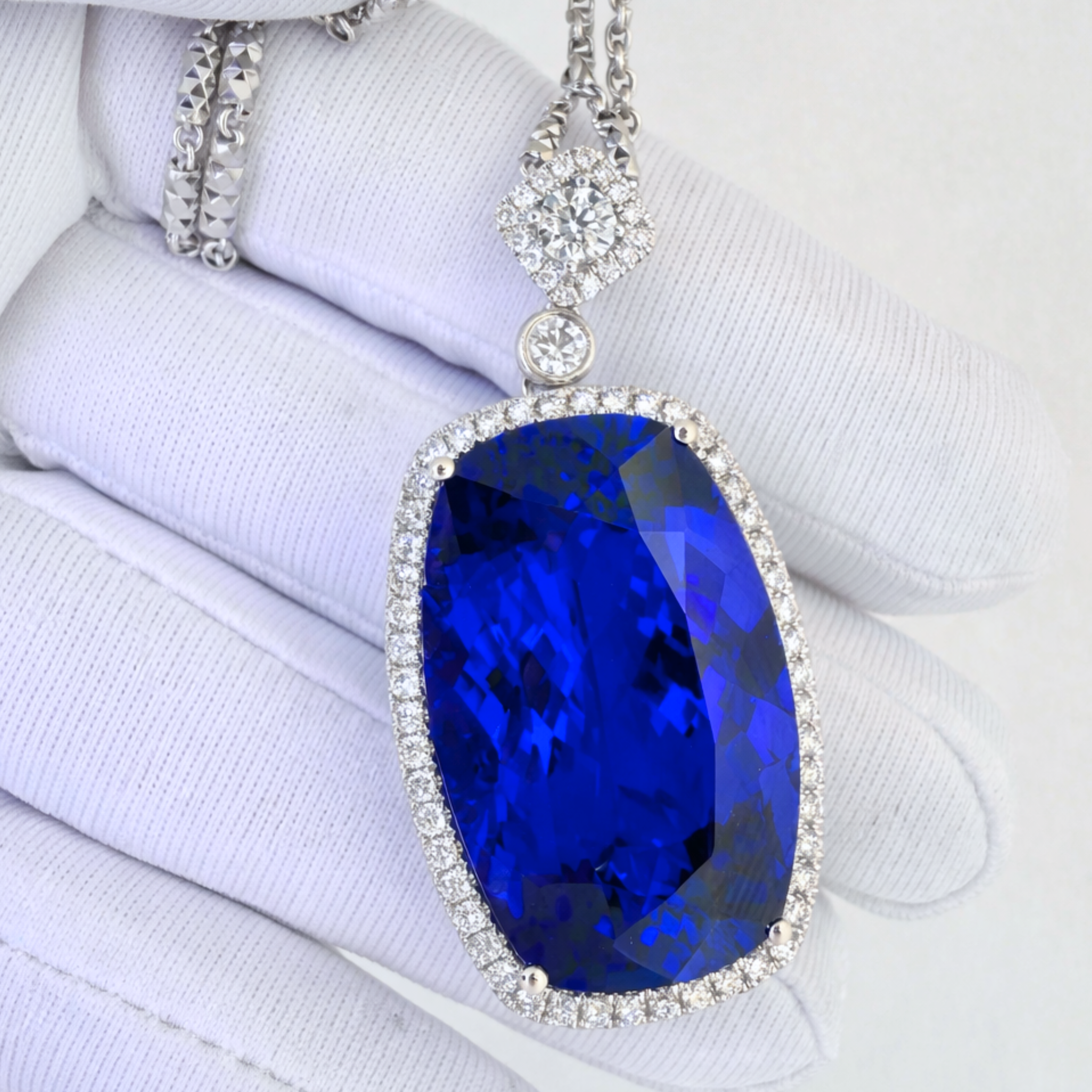 A large oval-cut vivid blue  tanzanite gemstone pendant with a silver-toned halo and chain, displayed on a white gloved hand.